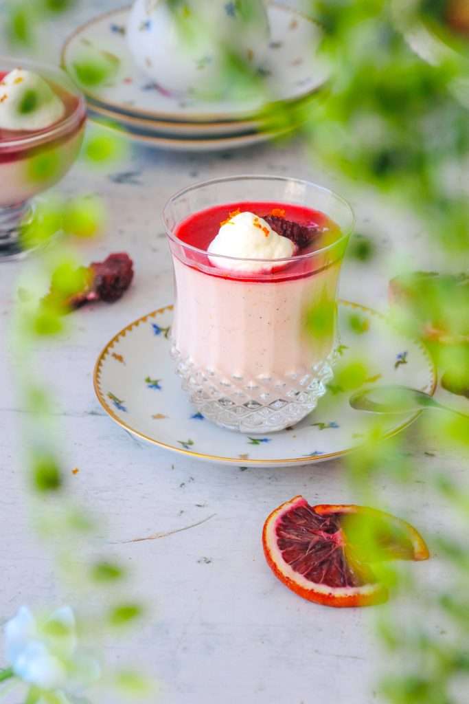 Blood orange panna cotta in a glass visible through green leaves.