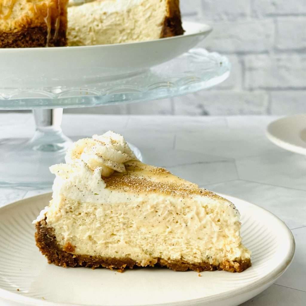 Nutmeg spiced cheesecake slice on a plate with the rest of the cake on a cakestand in the background.