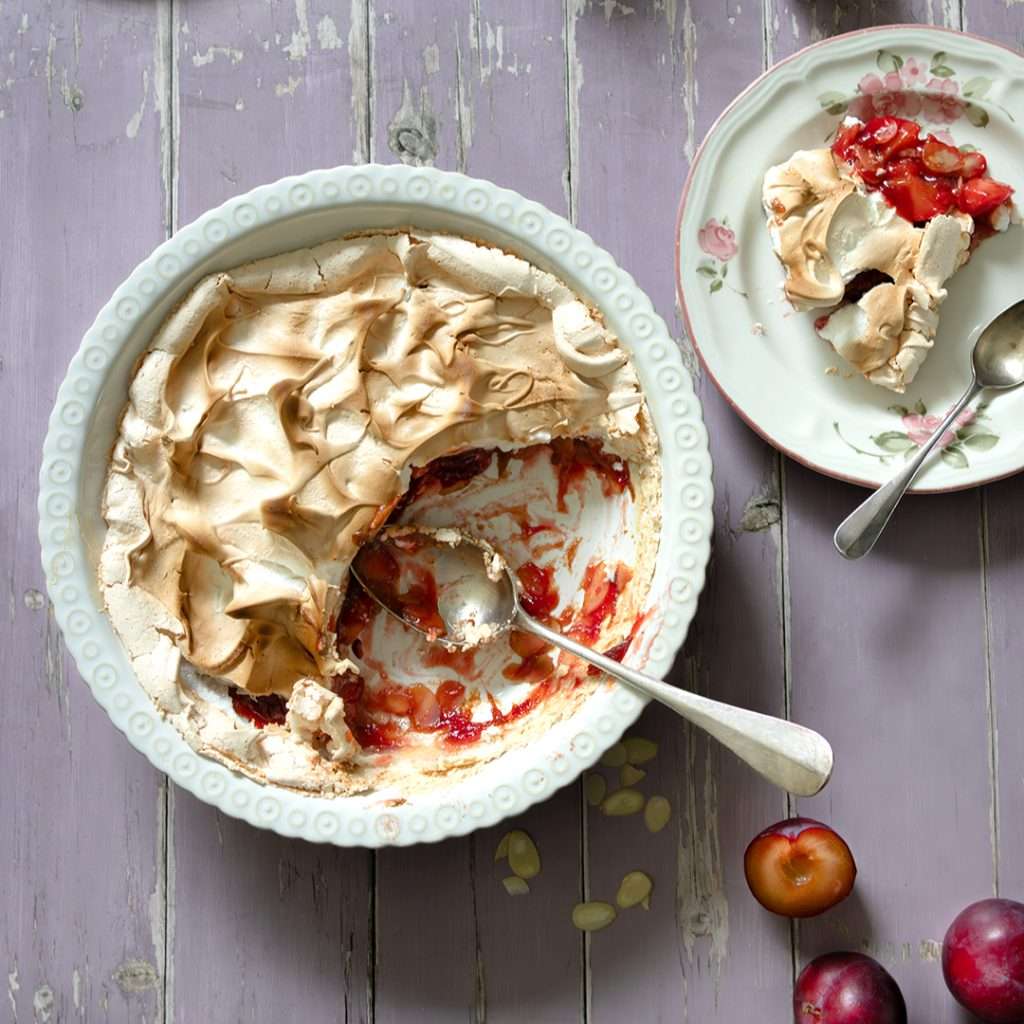 Swiss Plum Meringue (Zwetschgen) with Toasted Almond Base 1 Swiss plum meringue in a white circulsr baking dish with scoops taken out to show the layers of soft meringue and cooked plum filling.