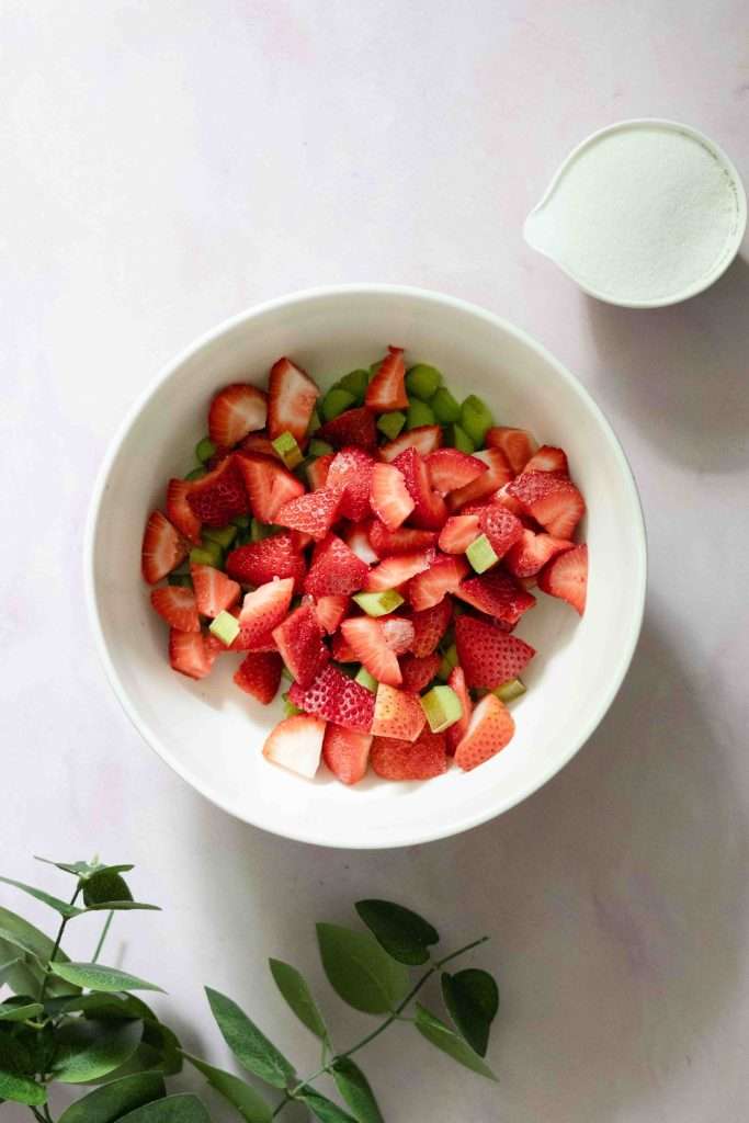 Cut strawberries and rhubarb in a white mixing bowl with a bowl of sugar besides it.